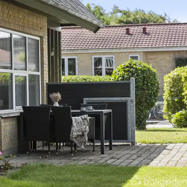 En terrasse med et bord og fire sorte stole står op ad en gul murstensbygning. Området er omgivet af grønne buske og en velplejet græsplæne.