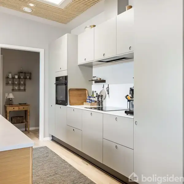 Køkken med hvide skabe og indbygget ovn i en moderne, minimalistisk stil. En bordplade med diverse køkkenredskaber, mens et tæppe løber langs gulvet. En dør fører til et andet rum.