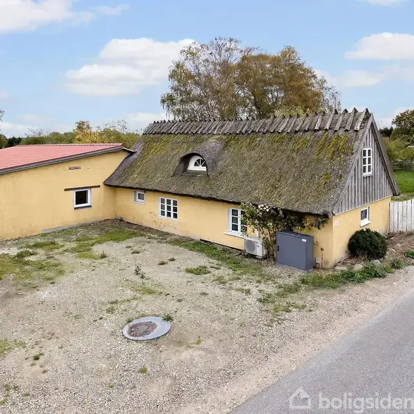 Gul, stråtækt bygning med en nyere tilbygning og en lille have foran, beliggende ved en landevej. Skyfri, blå himmel og enkelte træer i baggrunden.