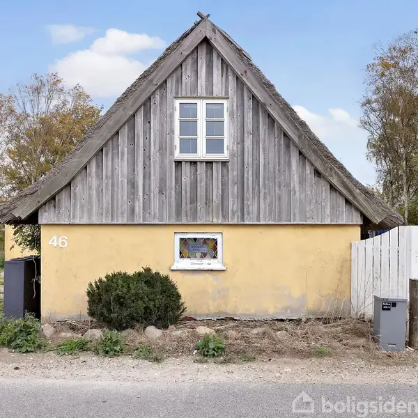Et ældre hus med stråtag står stille i et landligt område. Nummer 46 er malet på den gule facade. Foran er en lille busk og en hvid stakitlåge.