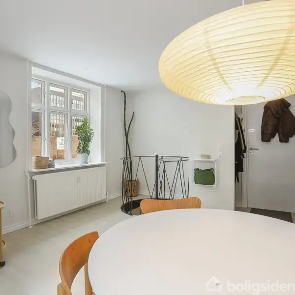 Rundt bord med træstole står i et lyst rum. En stor papirlampe hænger over bordet. Væggene er hvide, og der er et vindue samt en lille trappe i baggrunden.