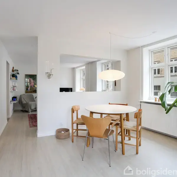 Et rundt bord med fire stole står i et lyst rum. En lampe hænger over bordet. Til højre er der et stort vindue og en plante.