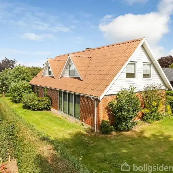 Et rødstenshus med rødt tegltag og to loftvinduer står stille i en velholdt have med grøn græsplæne og buske. Nabohuse ses i baggrunden under en blå himmel.