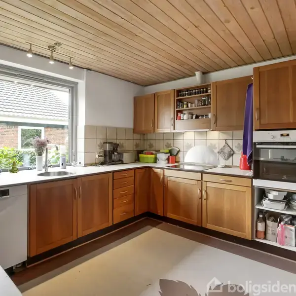 Køkken med træskabe og hvidevarer, inkluderer indbygget ovn og opvaskemaskine. Rummet har et stort vindue med udsigt til en have og er udstyret med lyse fliser og træloft.