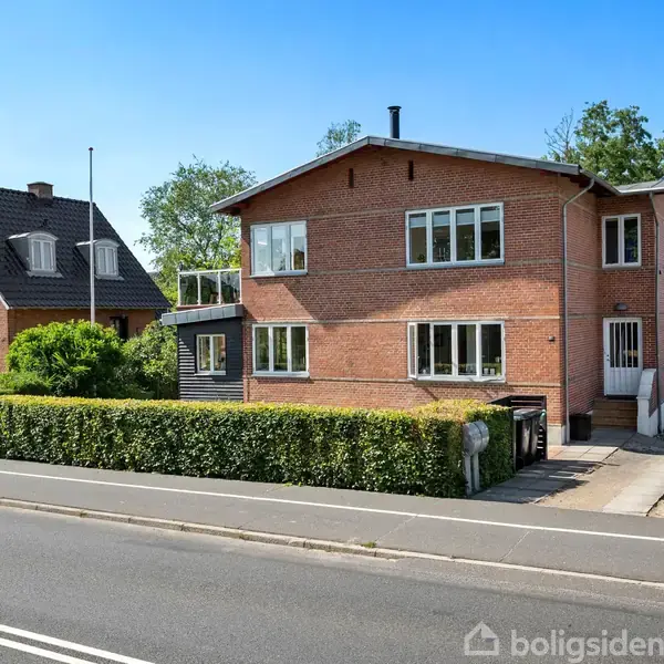 Rødt teglhus med hvide vinduer står ved en asfalteret vej. Huset har en indkørsel med en carport til højre. Grønt hegn omgiver grunden. Sollys skinner, og himlen er blå.