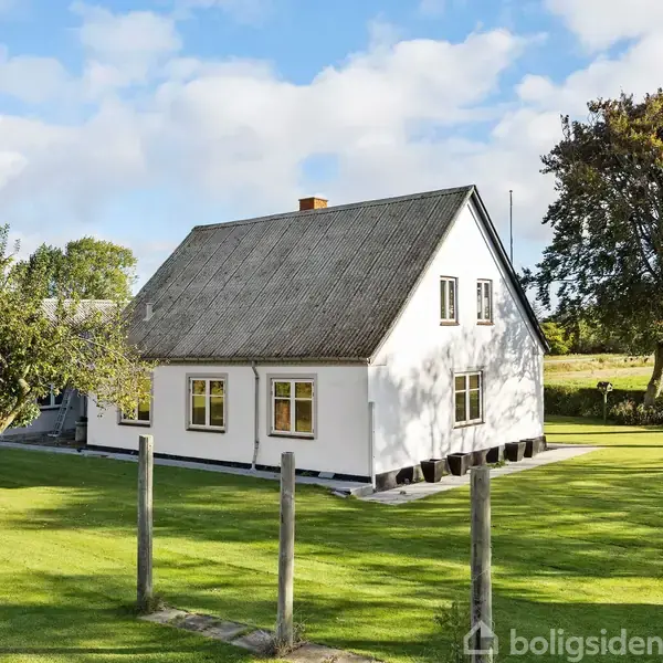 Hvide hus står i en grøn have med velholdt græsplæne og æbletræer. Det rummer vinduer og et skrånende tag. Landskabet består af marker og træer under en blå himmel.