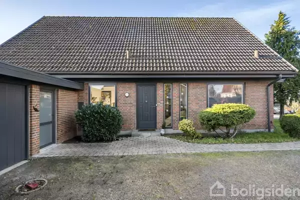 Et rødt murstenshus med skråt tag og hoveddør omgivet af buske. Der er en indgangssti foran huset. En garage er forbundet på venstre side.