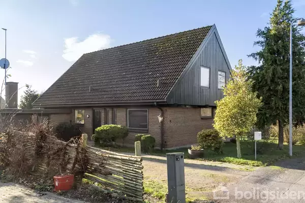 Et hus med mørkt skråtag står i en have med buske og træer. Foran er en ujævn grussti. Der er en postkasse ved indkørslen.