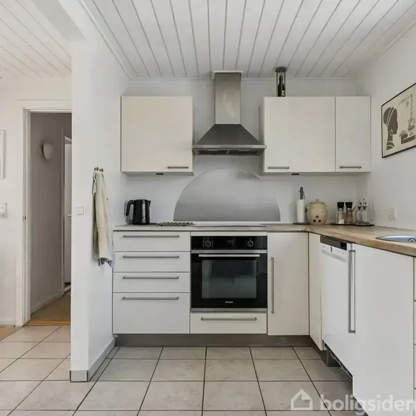 Køkken med hvide skabe og indbygget ovn, emhætte og vask. Flisegulv og træloft i et lyst rum. Malerier på væggen og planter på hylde.