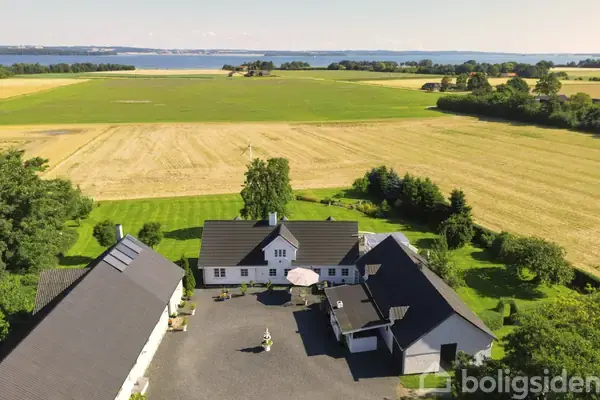 Landhus omgivet af marker, med havudsigt i baggrunden. Bygningerne står omkring en gårdsplads med grus, grønne træer, og marker strækker sig mod horisonten.