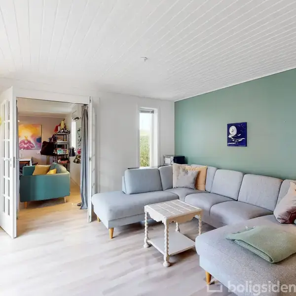 Grå sofa står i en lys stue med grønt malet væg bagved. Bord er placeret foran. Rummet har store vinduer og åben dør til et andet rum med farverig sofa.