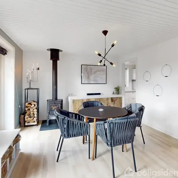 Et rundt bord med fem stole står i en lys spisestue. En moderne lysekrone hænger over bordet. Pejsen er placeret ved siden af en stabel brænde.