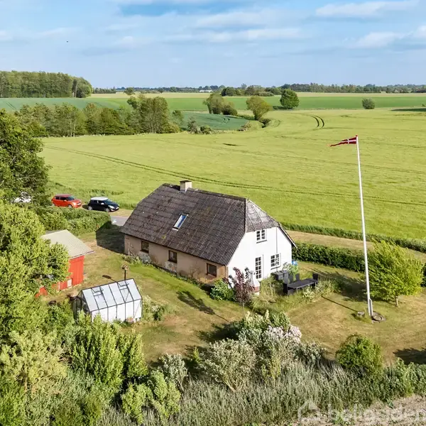 Et hvidt, stråtækt hus står stille omgivet af åbne marker og træer. Et flag vajer i haven nær en lille grøn bygning og et drivhus.