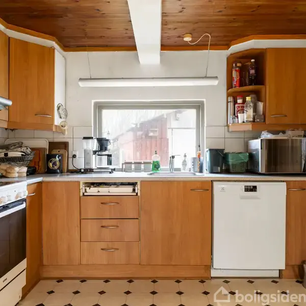 Et køkken viser trækøkkenskabe og skuffer med diverse køkkenmaskiner og krydderihylder. En opvaskemaskine og en ovn er synlige. Gennemsigtigt vindue med udsigt til udendørsområde.