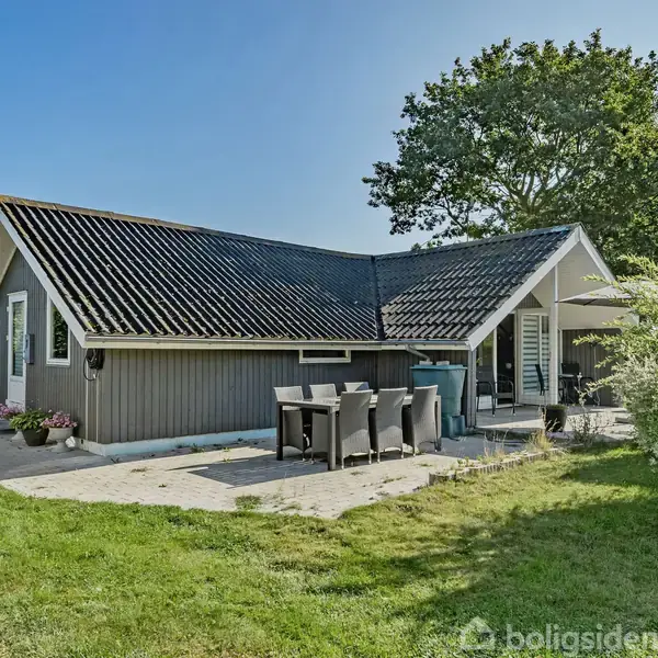 Gråmalet hus med sort tag står i en grøn have. Ved huset er en terrasse med bord og stole under åben himmel. Flere planter omgiver huset.