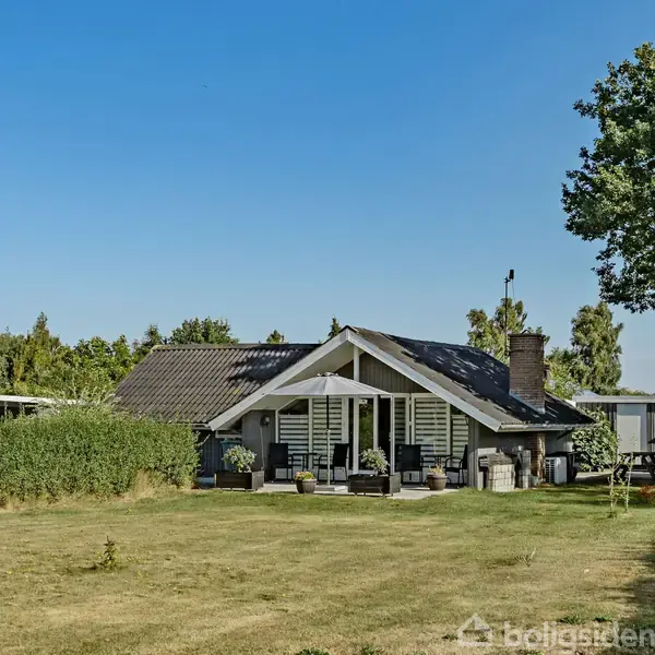 Hus med en terrasse udstyret med havemøbler, placeret på en stor græsplæne i et solrigt, grønt område med buske og træer i baggrunden.