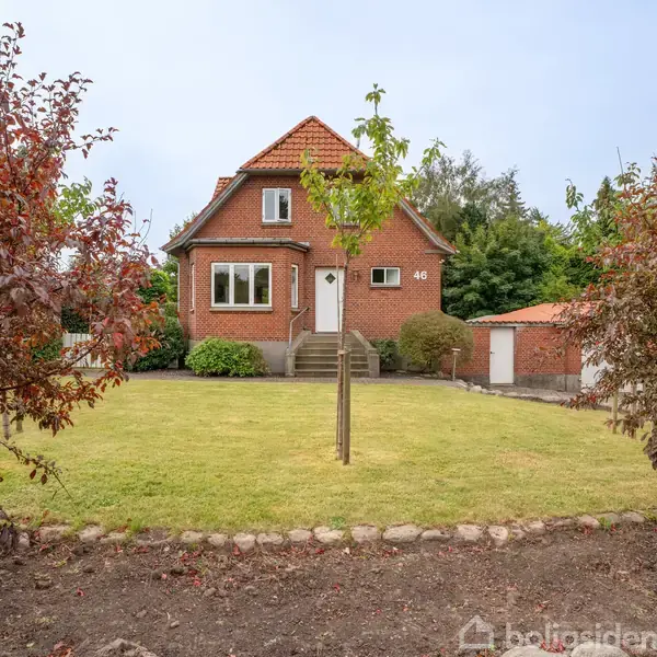 Et rødt murstenshus står stille i midten af en velholdt have omgivet af buske og træer. Huset har to etager med facadenummeret "46" synligt ved siden af døren.