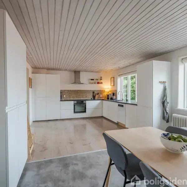 Et moderne køkken med hvide skabe står i hjørnet. Et spisebord med fire stole er opstillet i forgrunden. Store vinduer giver udsigt til grønne træer udenfor.