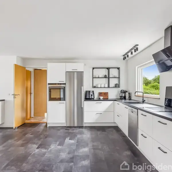 Køkkenet har hvide skabe og sort bordplade, midt i madlavning, omgivet af moderne apparater som ovn og køleskab. Dagslys strømmer ind gennem et vindue mod højre.