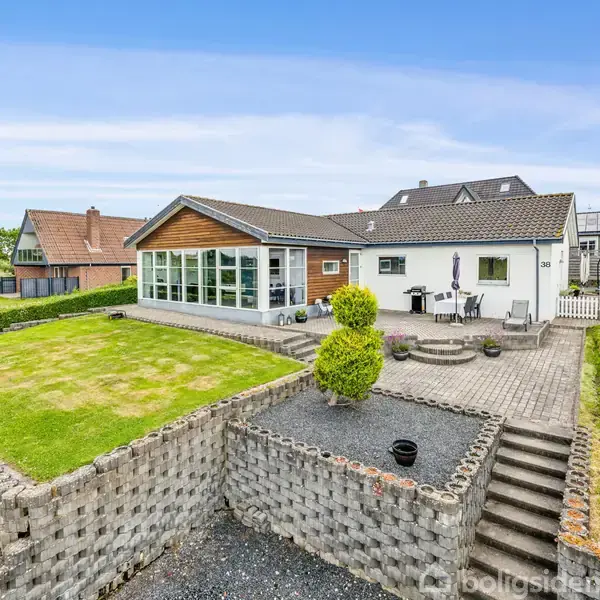 Et moderne hus med glasudestue står på en terrasse omgivet af en velplejet have med græsplæne og brolagte stier. Naboer kan ses i baggrunden, og himlen er klar.