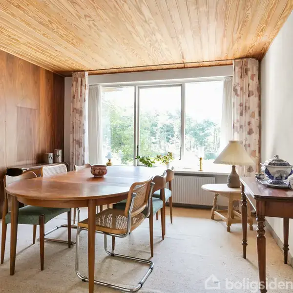 Rundt spisebord med stole står i et lyst rum ved et stort vindue med gardiner. En lampe og dekorative genstande er placeret på et konsolbord ved siden af.