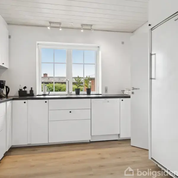 Et lyst køkken med hvide skabe og apparater. En kaffemaskine står på bordpladen ved siden af en brødrister. Store vinduer giver udsigt til en grøn have udenfor.