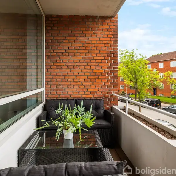 Et møbelarrangement med sorte lounge-sofaer og glasbord står på en altan. Der er en vase med grønne planter på bordet. Rød murstensbygning og træer ses i baggrunden.