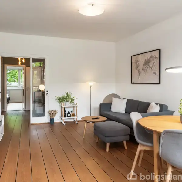 Grå sofa står i en stue med trægulv, ved et rundt spisebord med grå stole under en hængelampe. Vægdekoration inkluderer et kort. Planter optræder som dekoration.