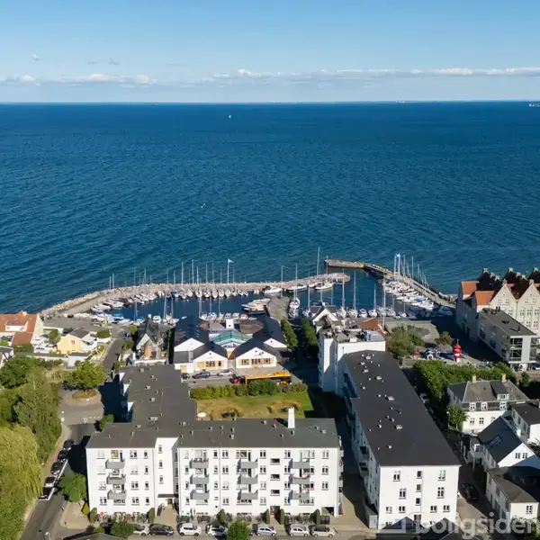 En lystbådehavn med mange både fortøjet i vandet. Baggrunden domineres af en dyb blå havudsigt. Forgrunden viser bygninger og boliger langs kysten.