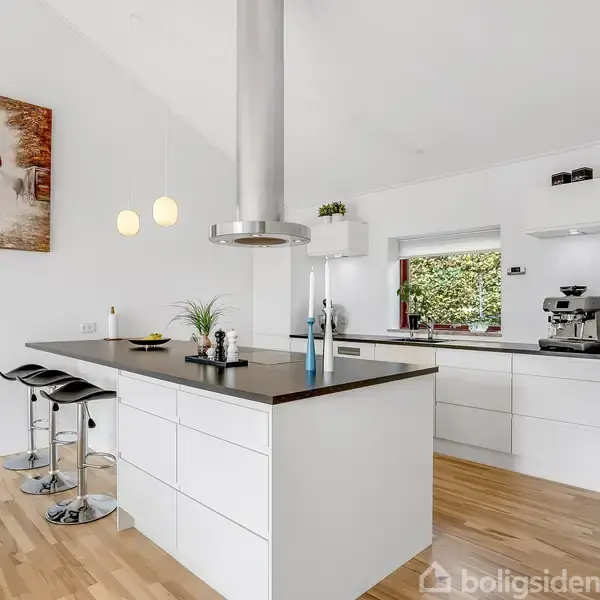 En terrasse med moderne udendørsmøbler står foran et murstenshus. Grå stole og et bord er omkranset af hække, grønne plæner og træer i baggrunden under en klar, blå himmel.