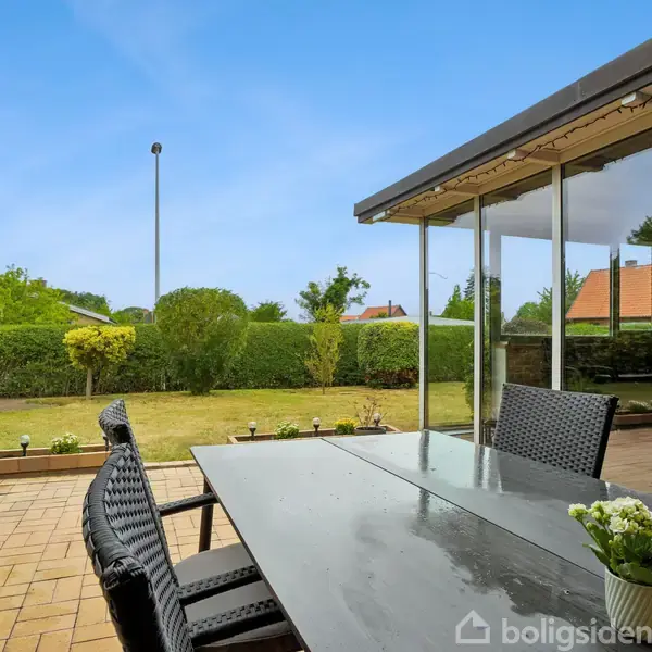Et udendørs spisebord med fire stole står på en terrasse. Parasol ved siden af. I baggrunden ses en velplejet have med græsplæne og buske under en klar blå himmel.