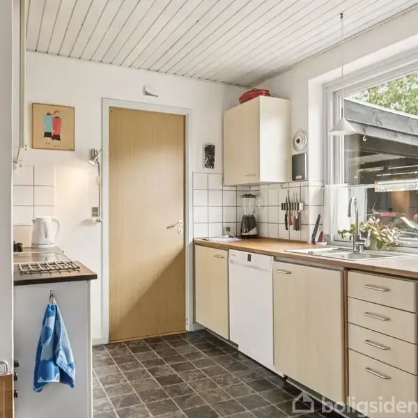 Et køkken med hvide skabe og skuffer, en stor vask under et vindue. Fliser på væggen, og en åben dør til en tilstødende gang. Tør håndklæde hænger på væggen.