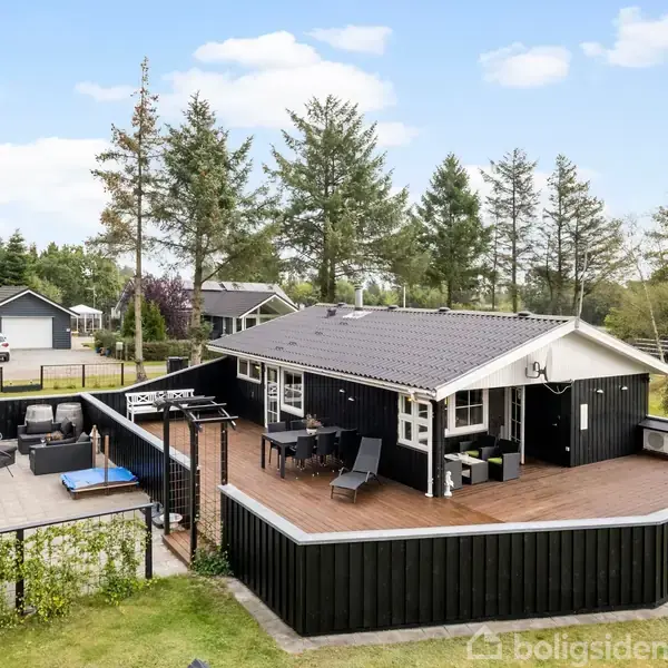 Sort sommerhus med stor træterrasse omgivet af hegn. Terrassen har udendørs siddepladser og står i en skovklædt område med flere træer i baggrunden under en klar blå himmel.