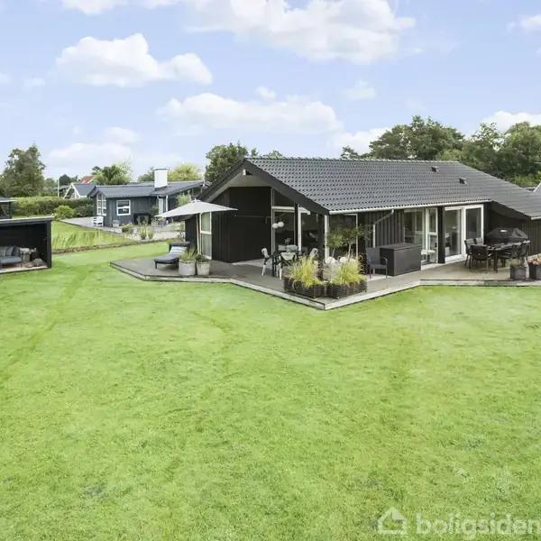 Et moderne sommerhus med stort terrasseområde står på en frodig græsplæne. En parasol giver skygge, og der er grønne planter omkring. Nabohuse ses i baggrunden sammen med træer og buske.