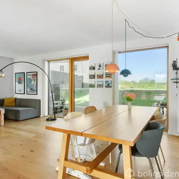 Et lyst, moderne stue med trægulv. En stor spisebord med stole er centralt placeret. I baggrunden står en hjørnesofa ved store vinduer og en glasdør, der slipper naturligt lys ind.