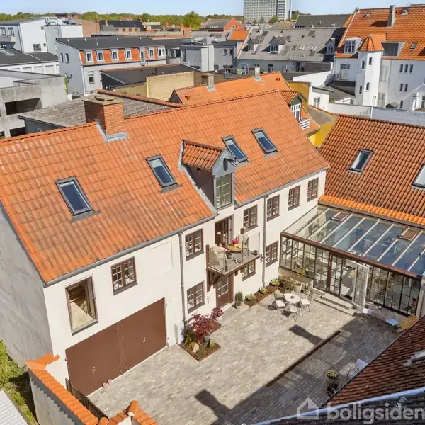 Et stort hus med rødt tegltag og flere ovenlysvinduer ses fra fugleperspektiv. Foran huset er en gårdhave med fliser og små borde. Ejendommen ligger i et tætbygd byområde.