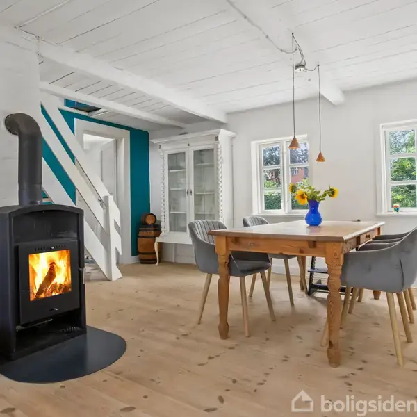 Brændeovn brænder ved en trævæg i en lys spisestue med træbord, stole og blomster i vase. Trætrappe og vinduer med udsigt til grønt.