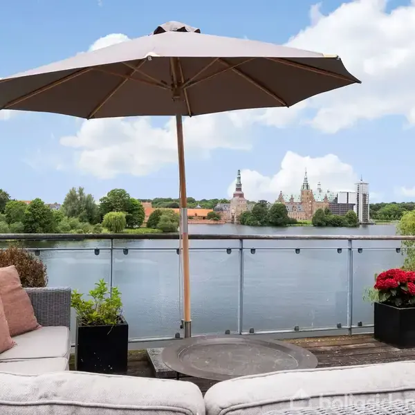 En terrasse med en stor parasol og hvide møbler ser ud over en sø med udsigt til en historisk bygning. Røde blomster tilfører farve til den grønne og blå omgivelse.