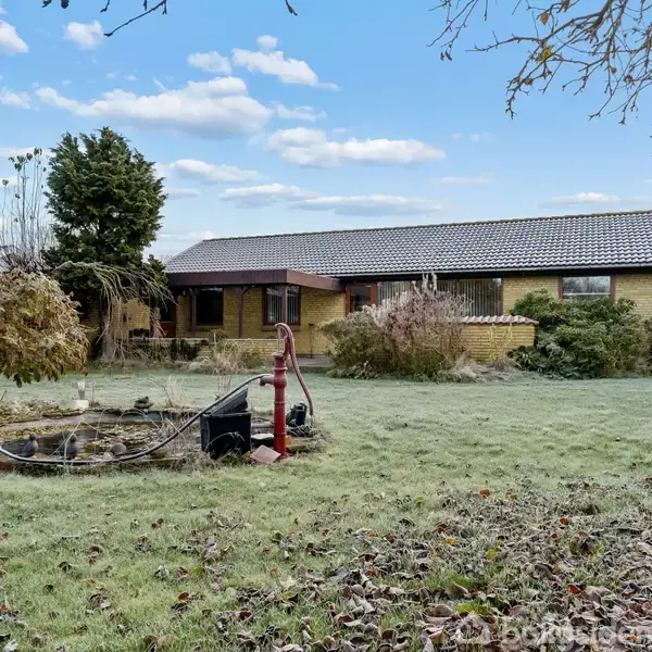 Hus med gult murværk og mørkt tag står i en have. En rød vandpumpe ved en lille dam omgivet af planter ses i forgrunden. Skyer på himlen.