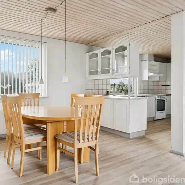 Et spisebord i lyst træ med fire stole står i et køkkenalrum med hvide skabe og flisebelagt væg. Store vinduer giver naturligt lys til rummet.