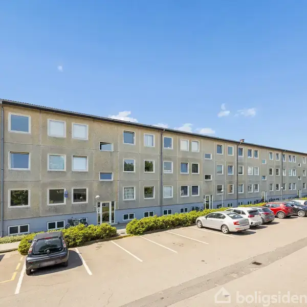 En grå murstensbygning med mange vinduer står langs en asfalteret parkeringsplads, hvor flere biler er parkeret. Bygningen er omgivet af grønne træer under en klar blå himmel.