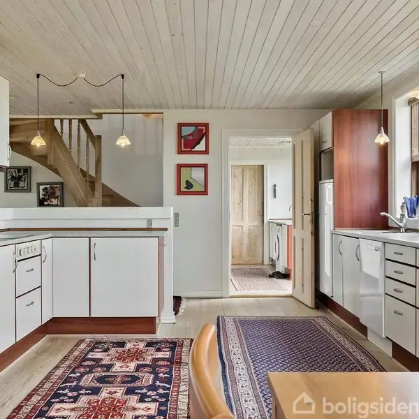 Køkkenet har hvide skabe og skuffer med trækantede håndtag, en indbygget ovn og hængende lamper. Det ligger i et hus med trægulve, tæpper og et lyst trætak.