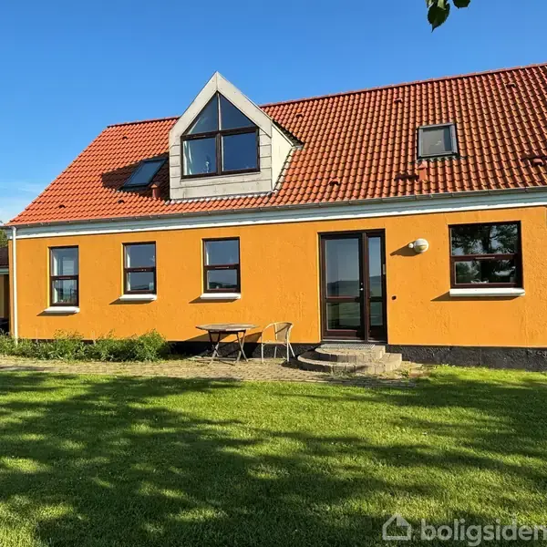 Et gult hus med rødt tegltag står på en grøn græsplæne. Huset har flere vinduer og en dør. Et lille bord med to stole er placeret udenfor.