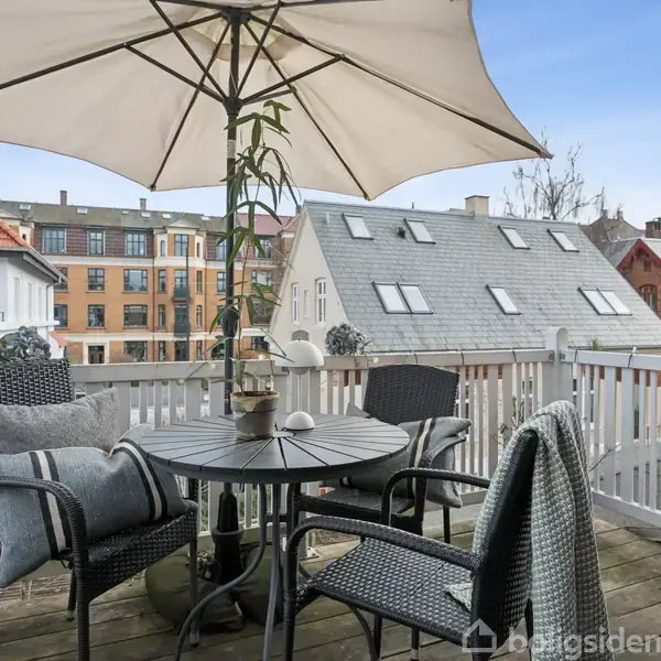 Terrasse med runde bord og tre stole, dækket af en stor parasol. Baggrunden viser hustage og bygninger i et bymiljø. Tæpper pryder stolene, og en plante står på bordet.