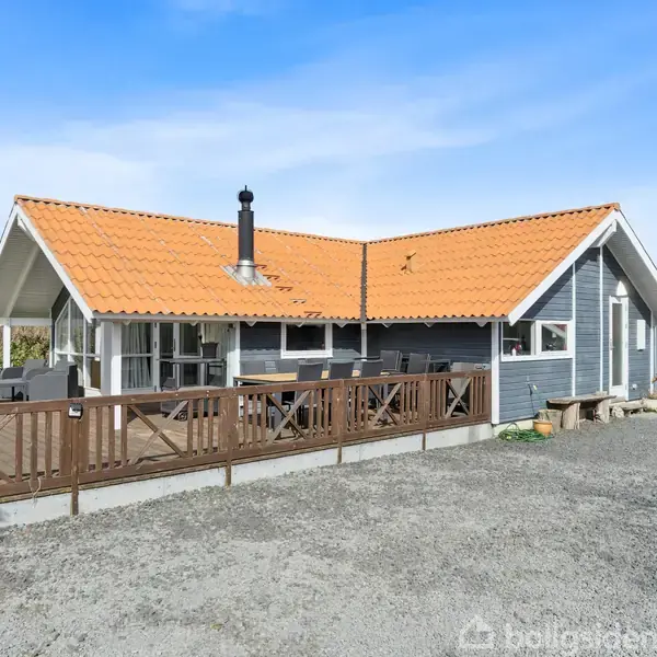 Et gråt træhus med orange tegltag står på en grusbelagt gårdsplads. Huset har en stor træterrasse med møbler og ligger omgivet af en grøn græsplæne og træer under en blå himmel.