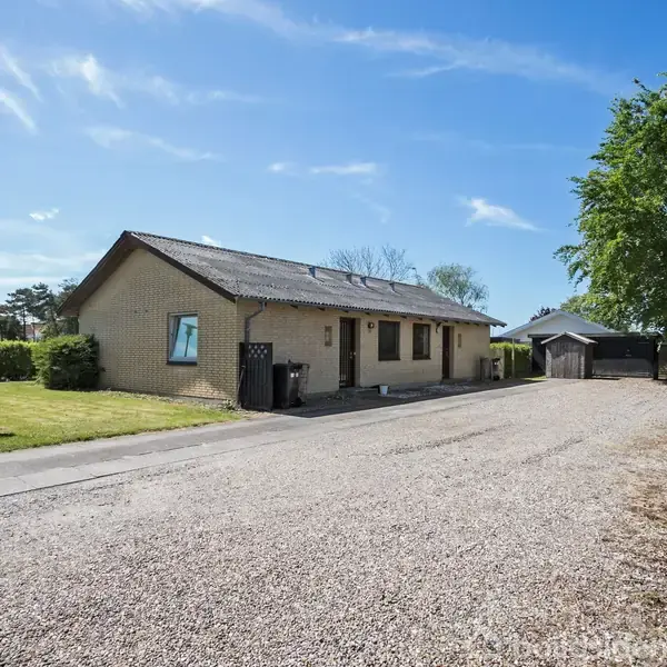 En enkeltstående gul murstensbygning står stille langs en grusvej, omgivet af grønne buske og træer, med en klar blå himmel i baggrunden.