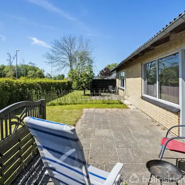 Solstol står ved siden af en rød stol på en flisebelagt terrasse. Græsplæne strækker sig til en hæk. Murstenshus med store vinduer er til højre. Klar blå himmel.