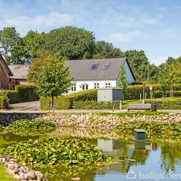 Et hvidt hus med tagvinduer står ved siden af en stenbelagt dam med åkander. Omkring ligger en vej, grønne buske, andre huse og træer under en blå himmel.