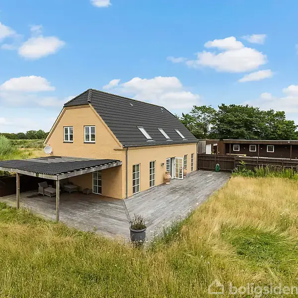 Et gult hus står stille omgivet af højt græs og træer, med en stor terrasse og en carport i en åben landlig omgivelser under en skyfri himmel.