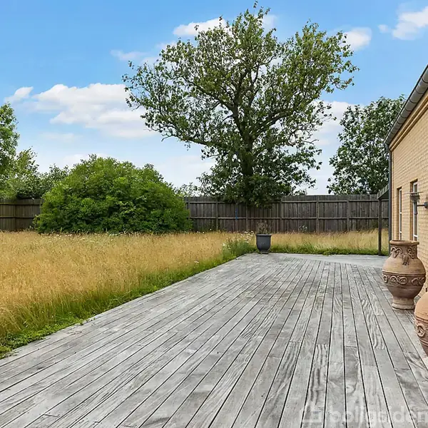 En træterrasse strækker sig langs en gul murstensbygning med store krukker i forgrunden. Højt græs og buske omgiver terrassen, omgivet af et plankeværk, under en blå himmel.